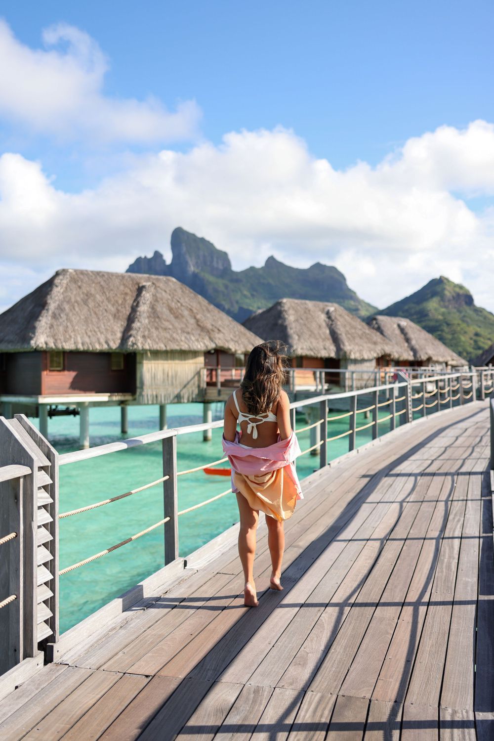 Overwater bungalows with wooden walkway against dramatic mountain backdrop in tropical paradise.