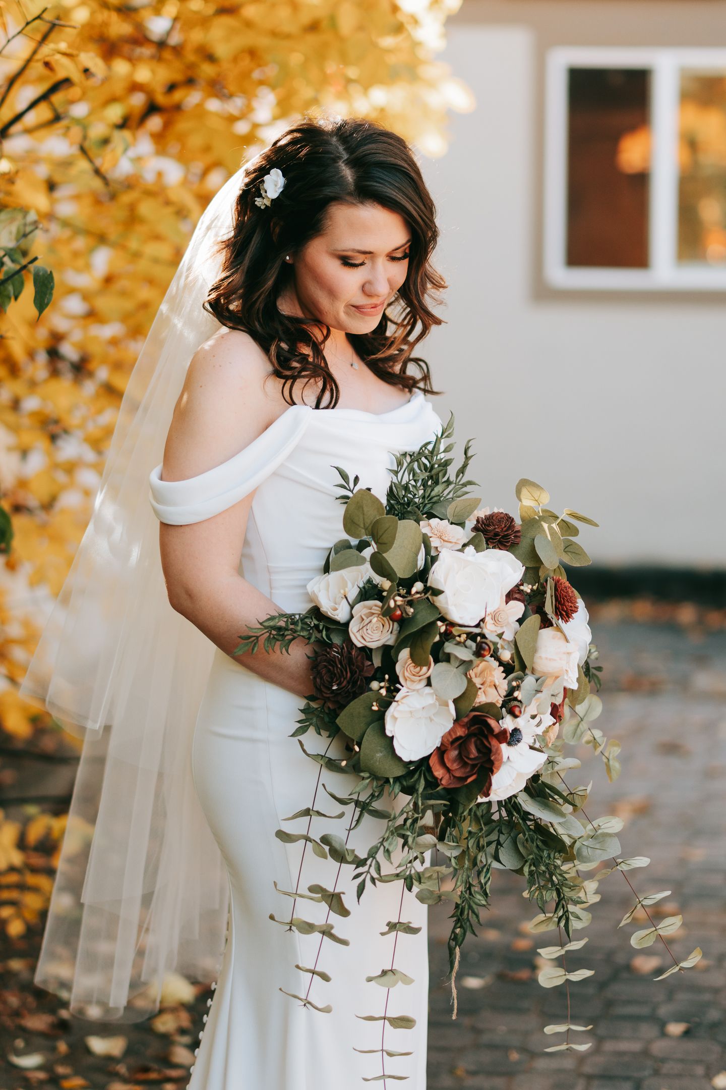 Bride in white dress holding elegant bouquet with autumn colors in background.