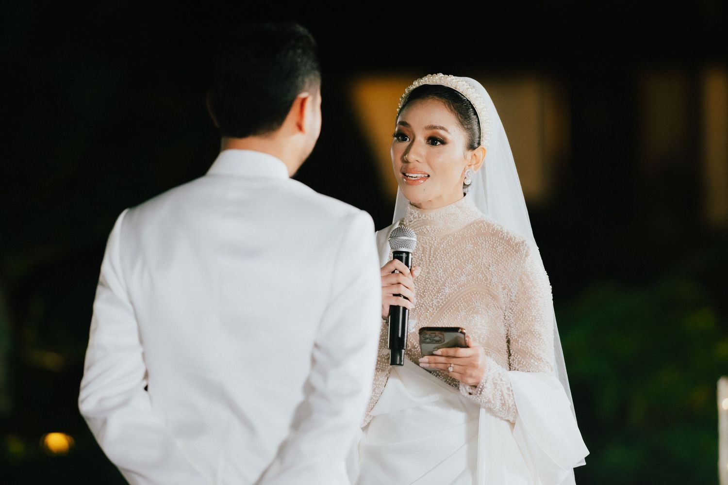 Bride in a beaded white gown and veil reading vows into a microphone at an outdoor night ceremony.