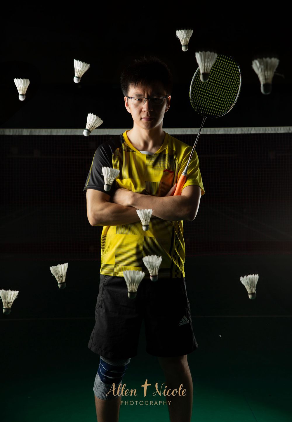Badminton player in yellow shirt stands with multiple shuttlecocks floating in dark studio setting.