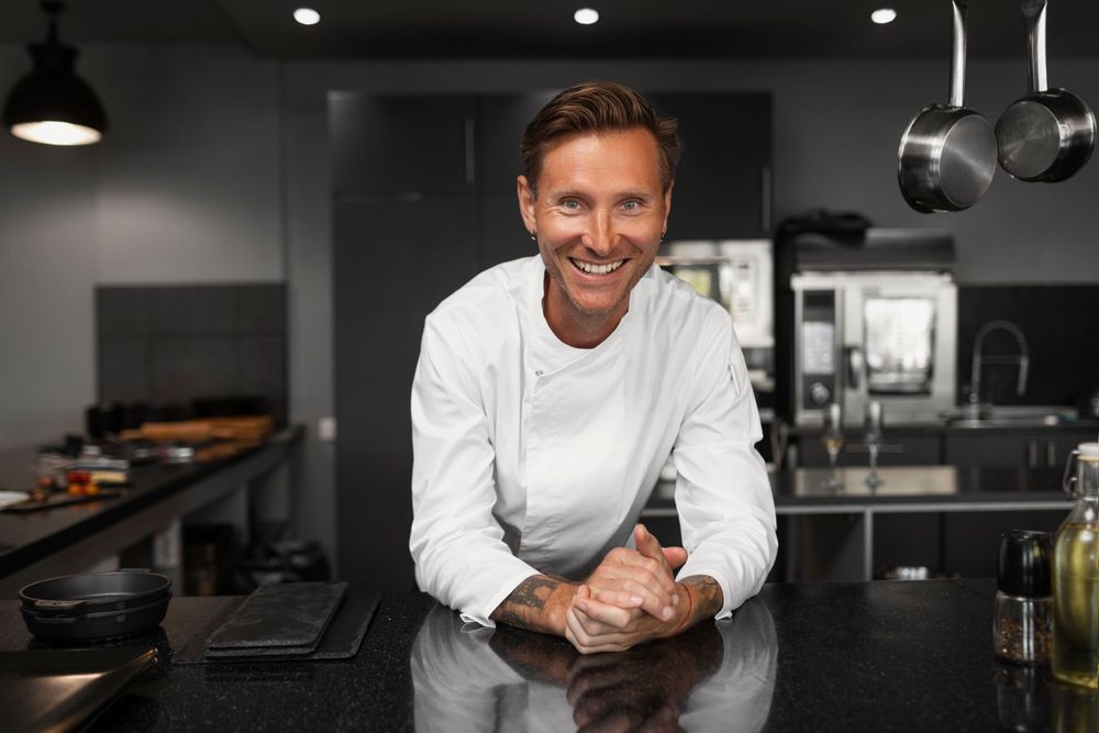 Professional chef in white uniform standing confidently in modern dark kitchen.