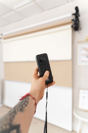 A tattooed hand holds a remote control aimed at a white projector screen mounted on a wall.