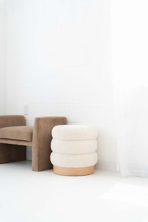 A minimalist room featuring a brown velvet chair and cream-colored round ottoman with wooden base against white walls.