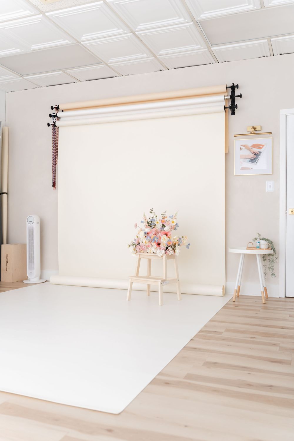 Clean white photography studio setup with neutral backdrop, wood floors, and delicate floral arrangement on wooden stool.