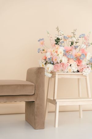 A pastel floral arrangement sits atop a white step stool next to a beige velvet sofa against neutral wall.