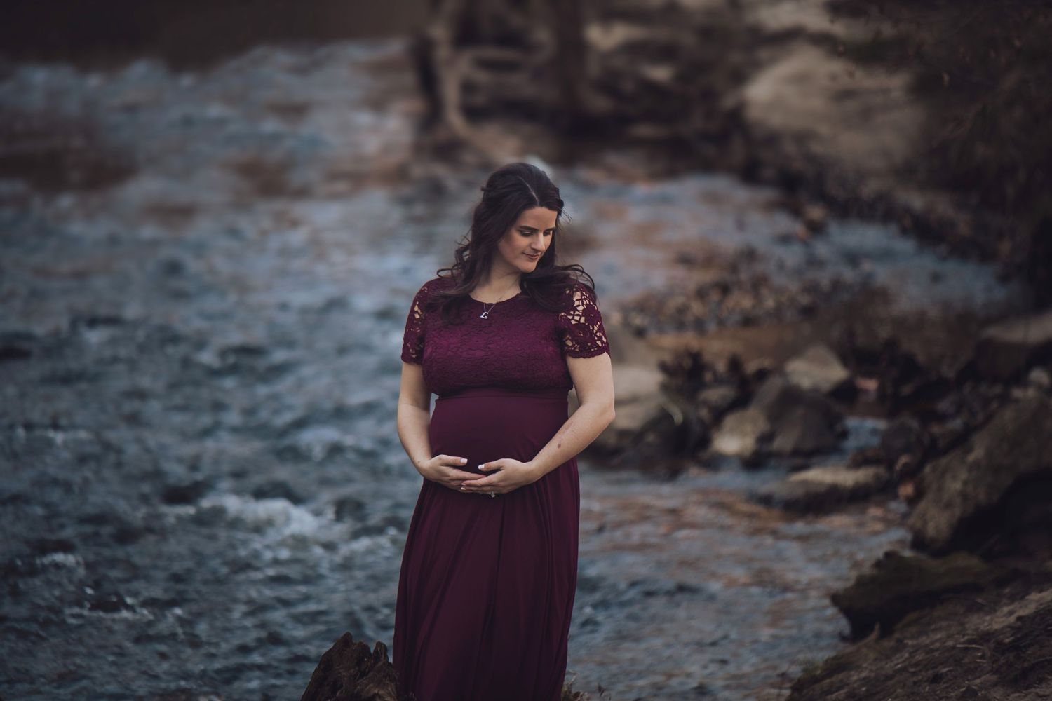 A person in burgundy dress standing near a rocky stream at dusk.