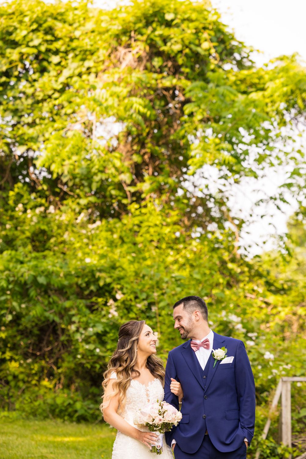 Wedding couple laughs together in a sunlit garden setting with lush green trees and foliage in the background.