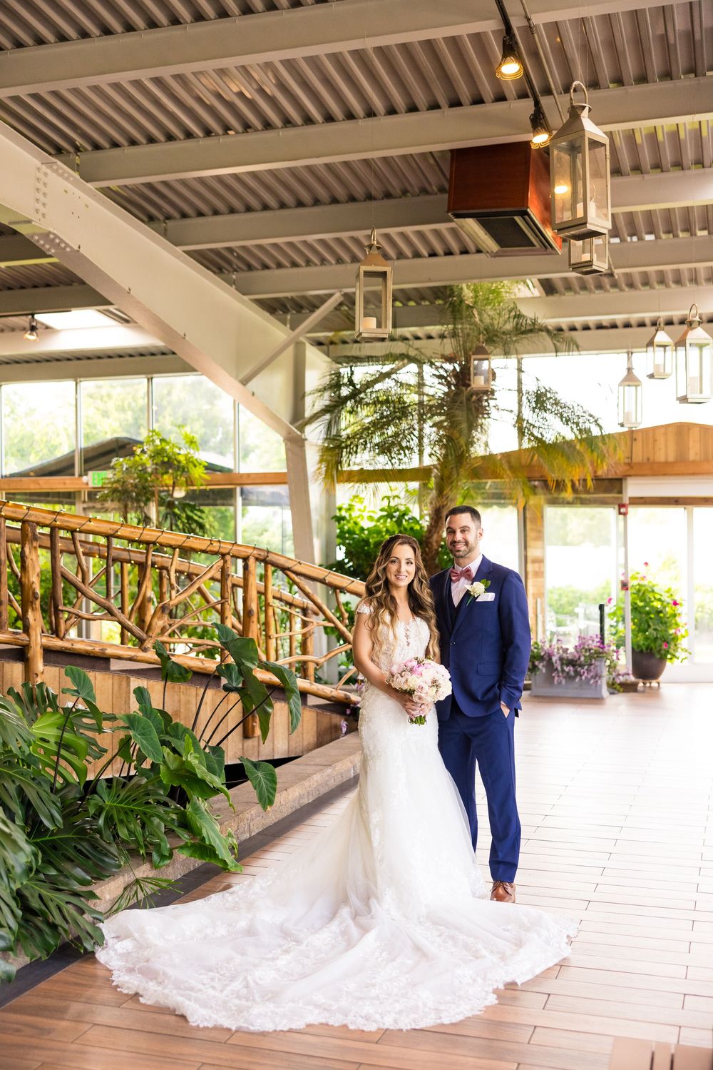 A couple poses at a beautiful greenhouse venue with ornate wooden railings and lush tropical plants surrounding them.