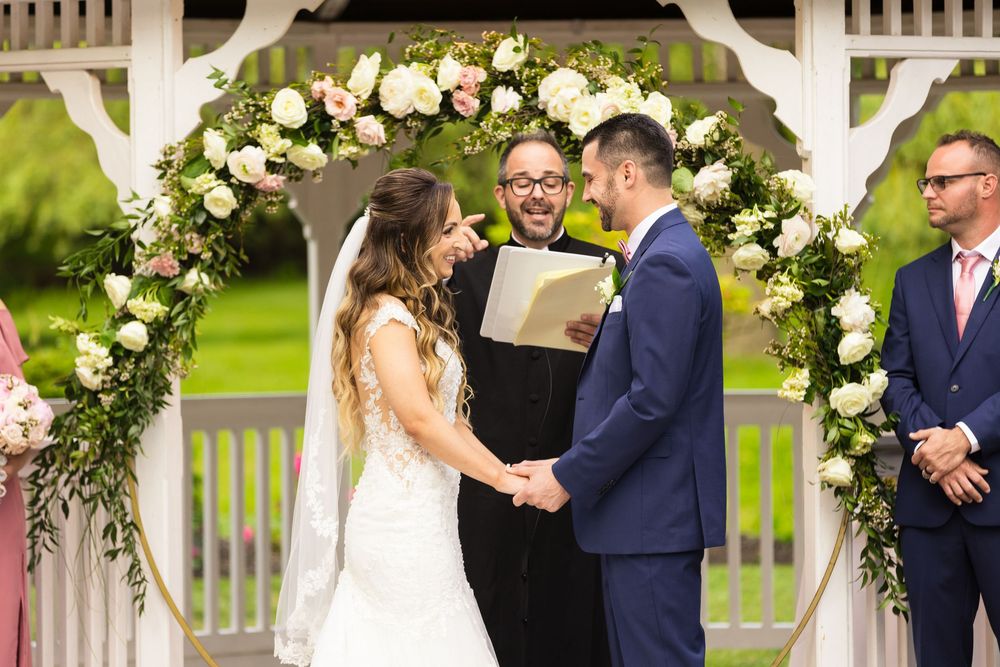 Wedding ceremony couple standing beneath floral arch decorated with white roses and greenery during outdoor celebration.