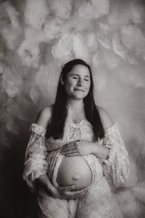 Black and white maternity photo of a smiling pregnant woman in lace dress cradling her baby bump against a floral backdrop.