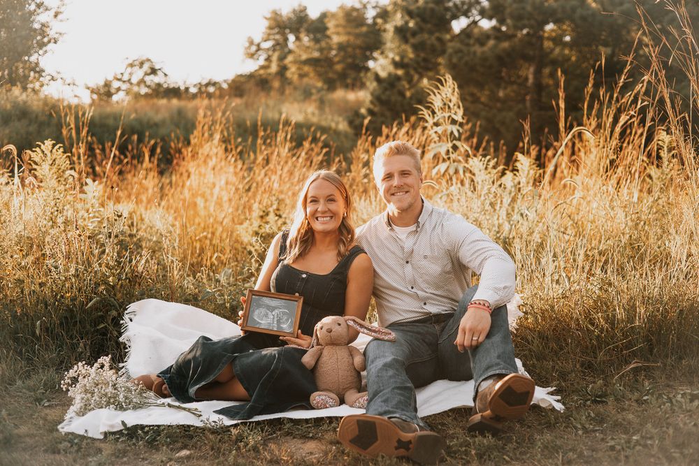 Happy couple sitting in golden field holding ultrasound photo and teddy bear for maternity announcement.