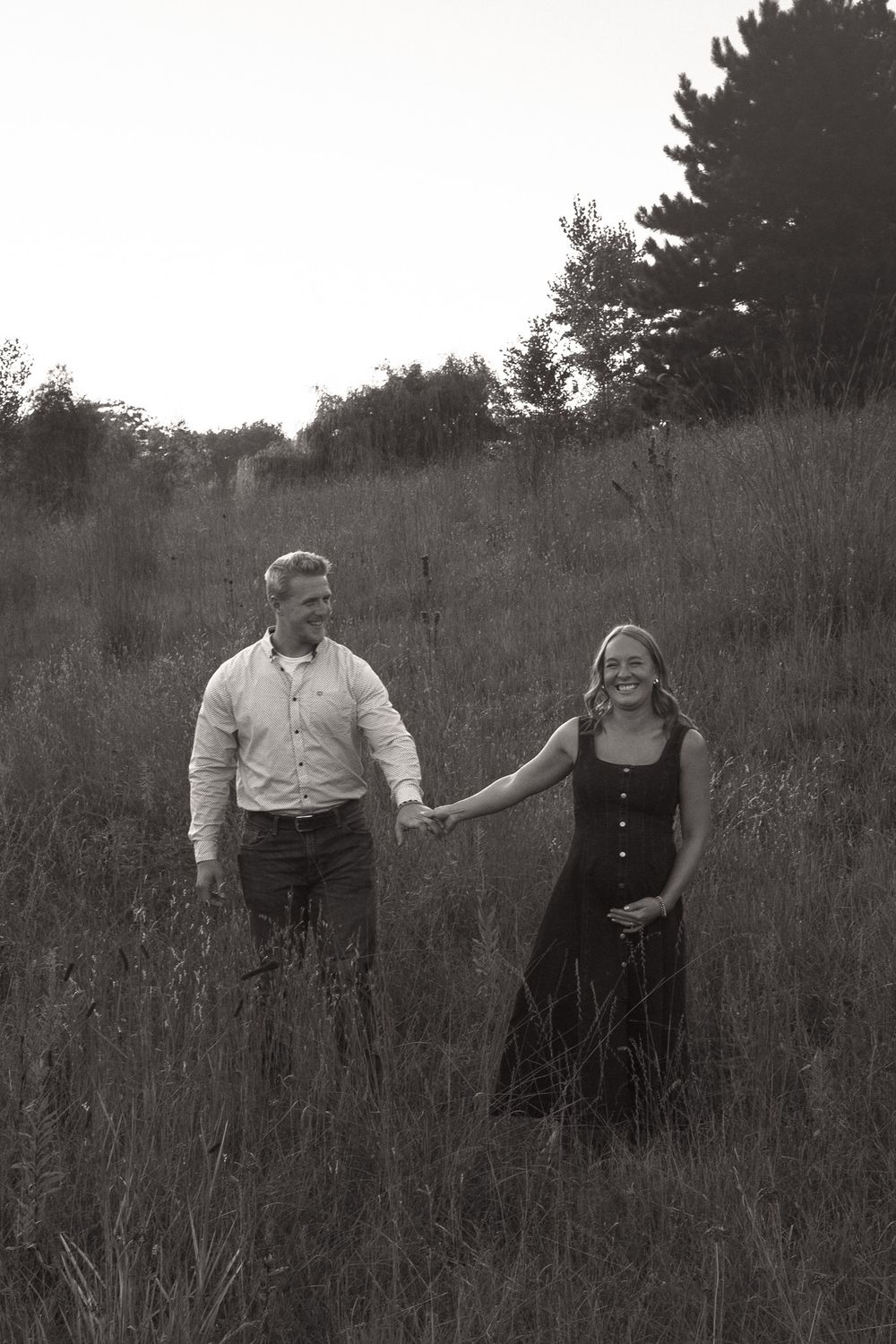 Black and white photo of a smiling couple holding hands while walking through a tall grass field with trees in the background.