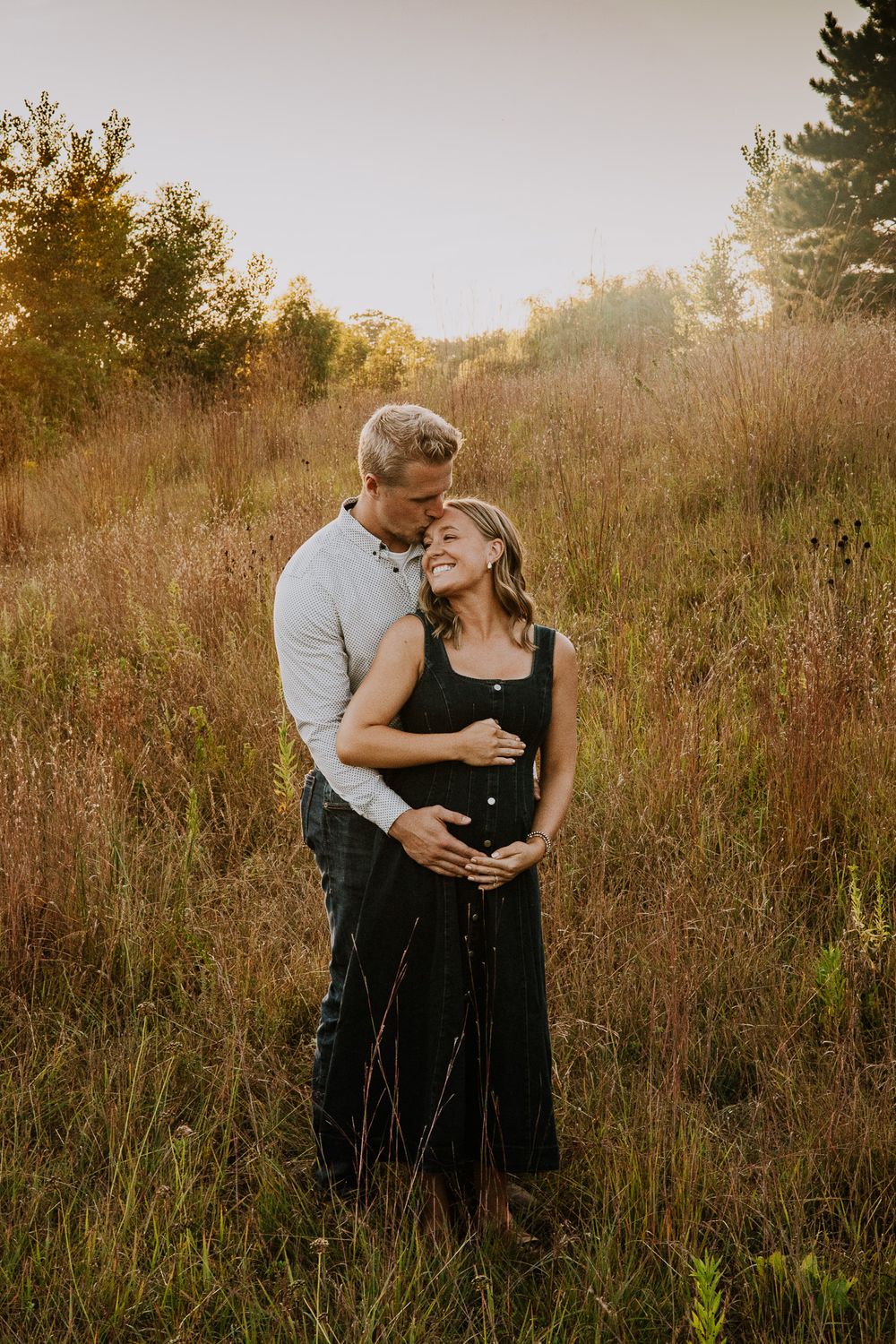 Couple embracing in golden meadow during sunset, pregnant woman in dark dress with partner holding her belly.