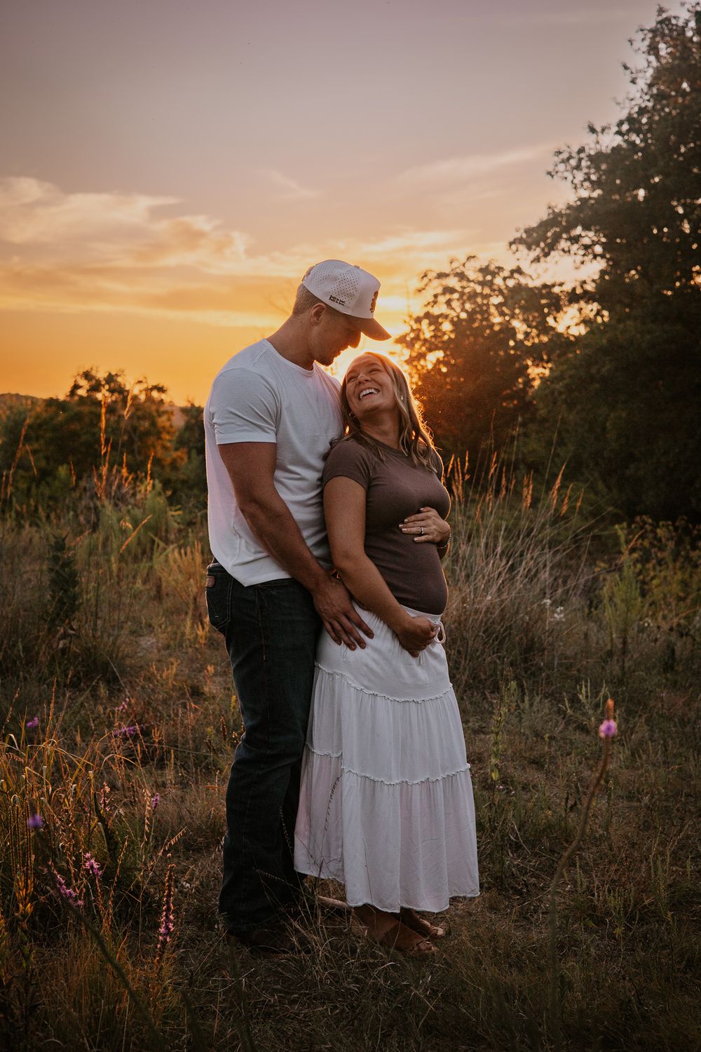 Couple embracing and laughing together in a golden sunset field, woman in white skirt touching her baby bump.