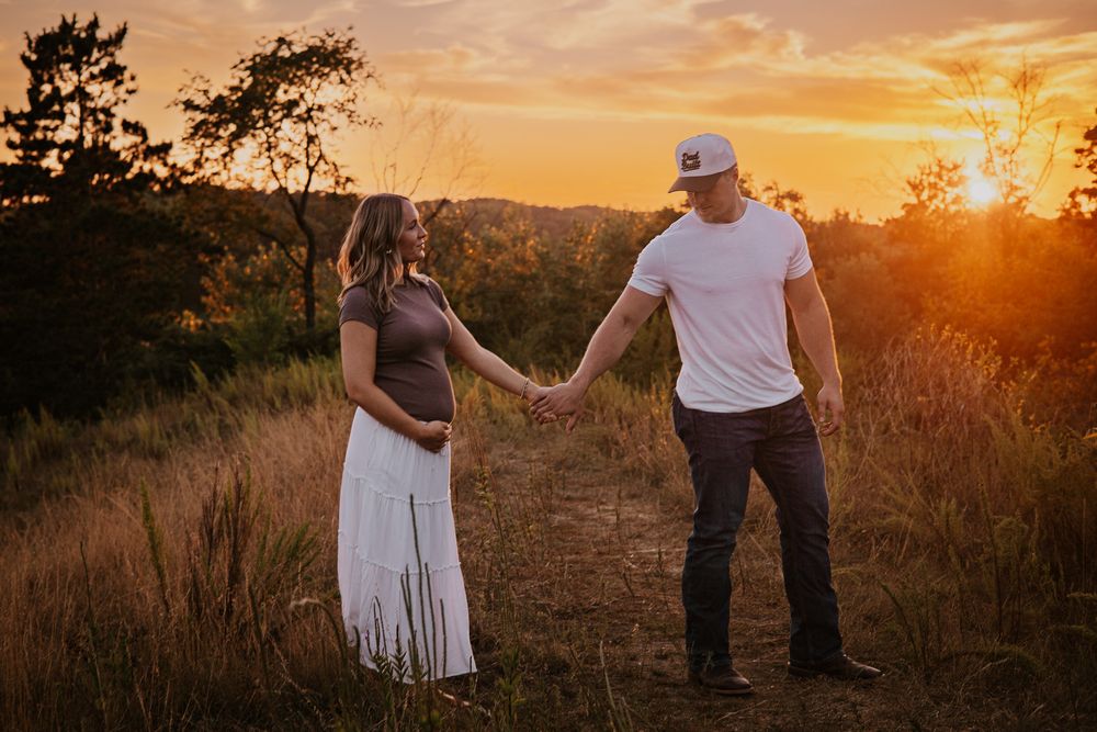 Pregnant couple holding hands in a golden sunset field, man in white tee and cap, woman in flowing white skirt.