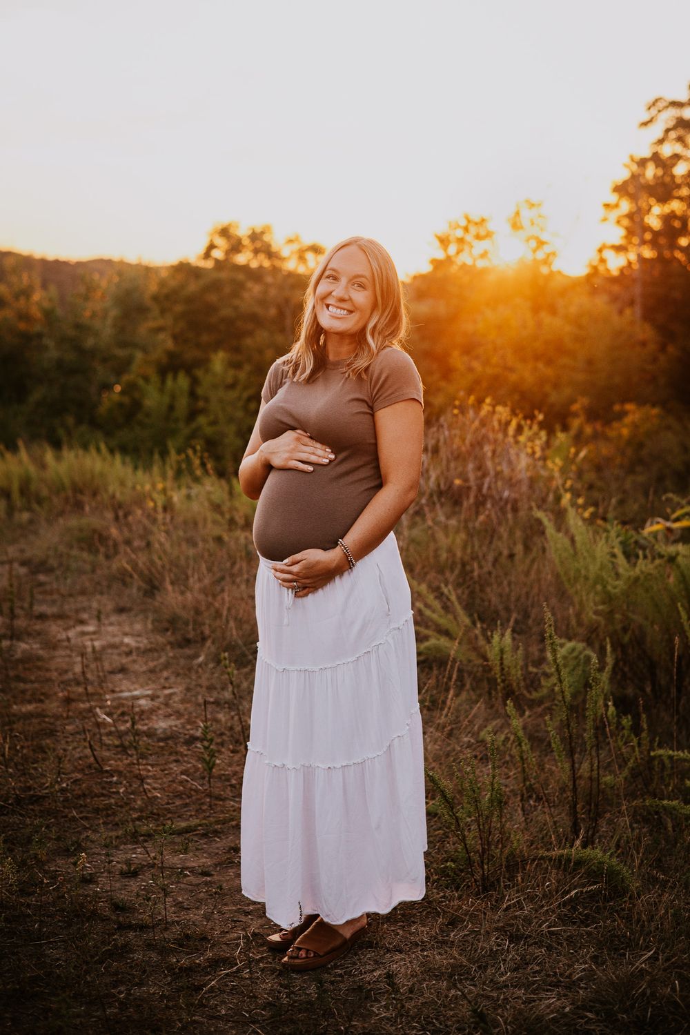 Smiling pregnant woman in brown top and white maxi skirt cradling baby bump at golden hour in nature.