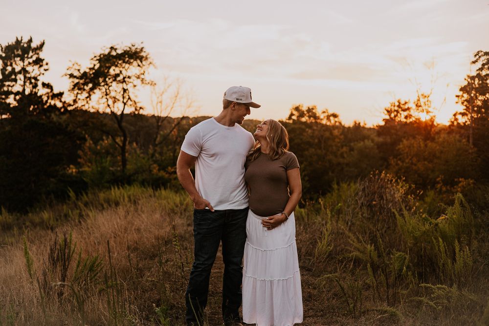 Couple sharing a tender moment at golden hour in a field, woman in white skirt cradling baby bump.