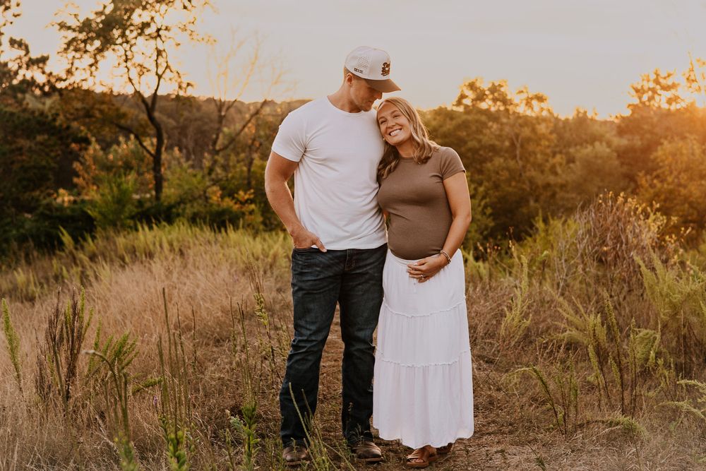 Couple posing outdoors at golden hour, pregnant woman in white skirt and man in white tee, surrounded by lush greenery.