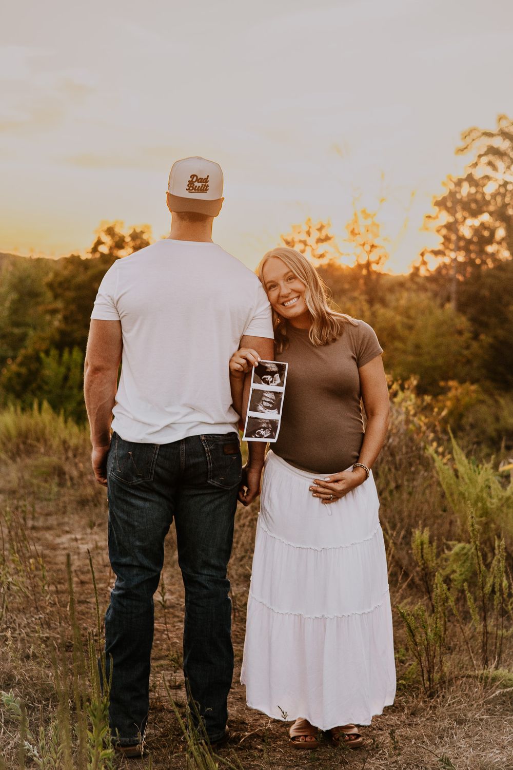 Pregnant couple holding ultrasound photos at golden hour outdoors, woman smiling in brown top and white maxi skirt.