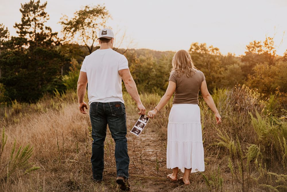 Couple walking hand in hand through golden field at sunset, holding ultrasound photos, pregnancy announcement outdoors.