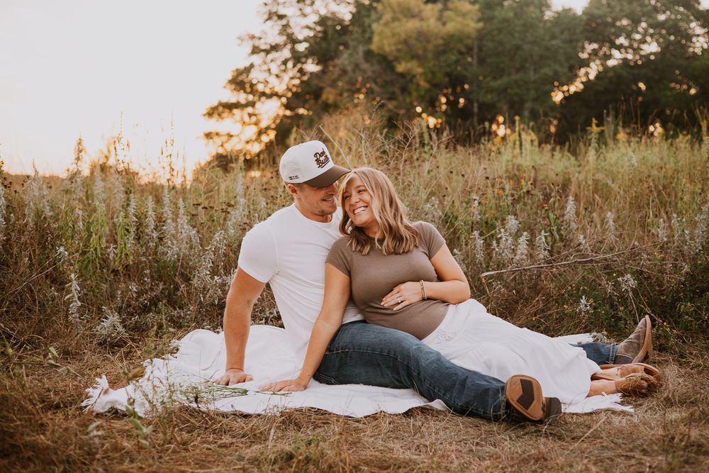 Couple sitting on white blanket in golden field, pregnant woman leaning on partner during maternity photo session.
