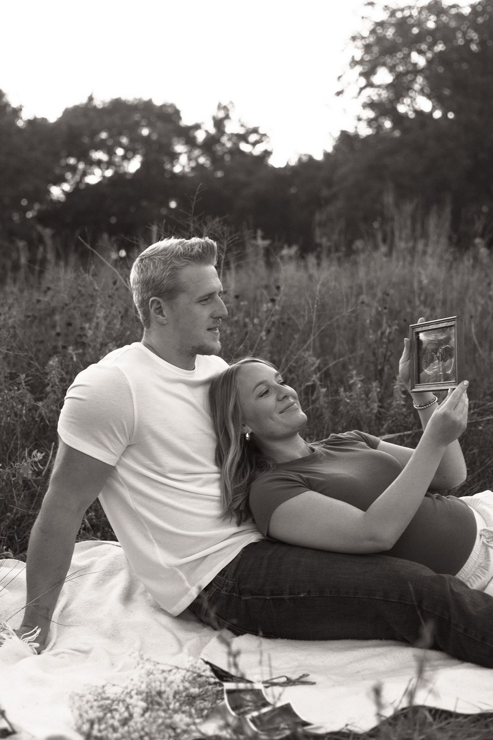 Black and white photo of a couple relaxing on a blanket outdoors, woman holding an ultrasound photo.