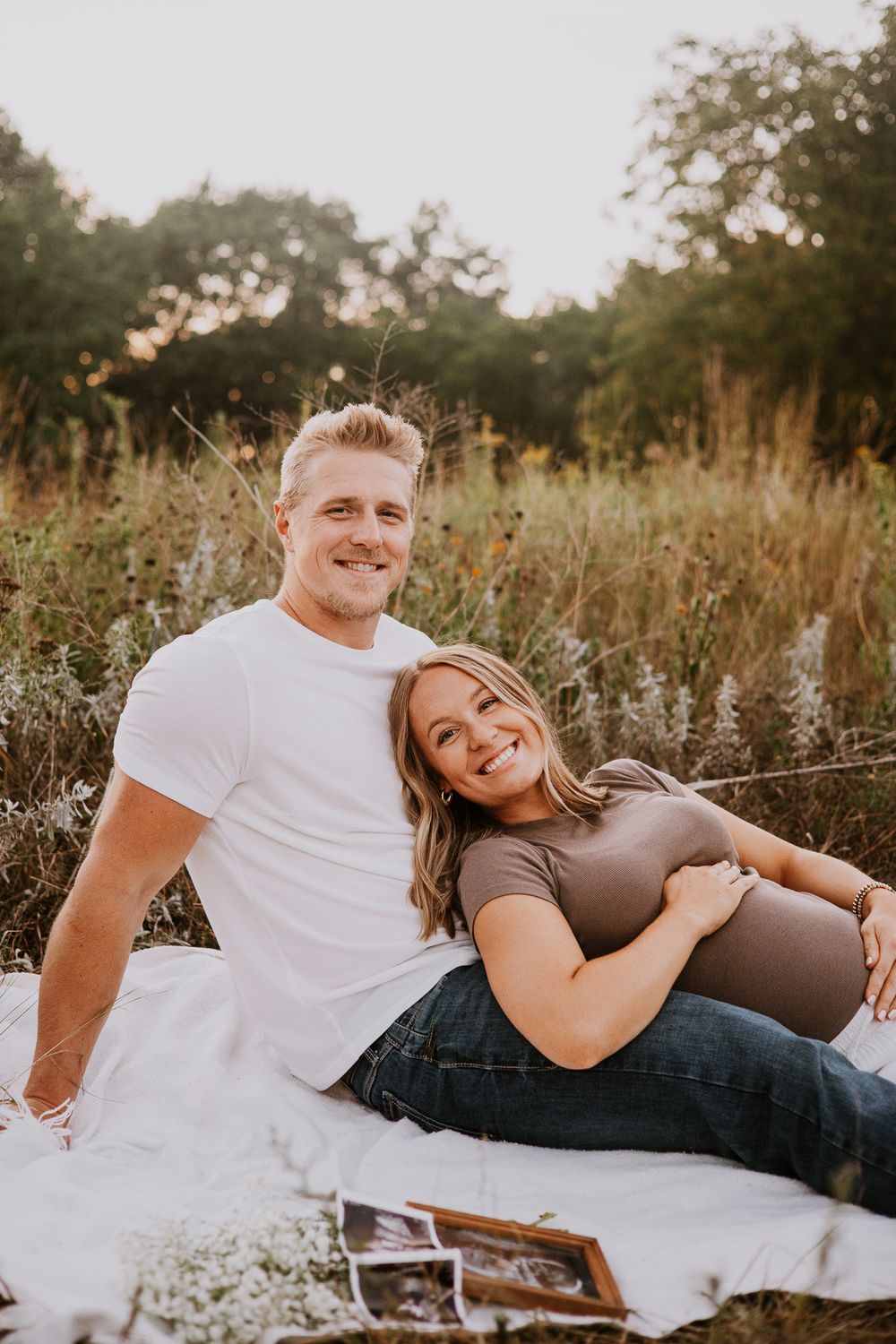 Happy couple sitting on a blanket in a field, woman resting on man's lap with baby bump and ultrasound photos.