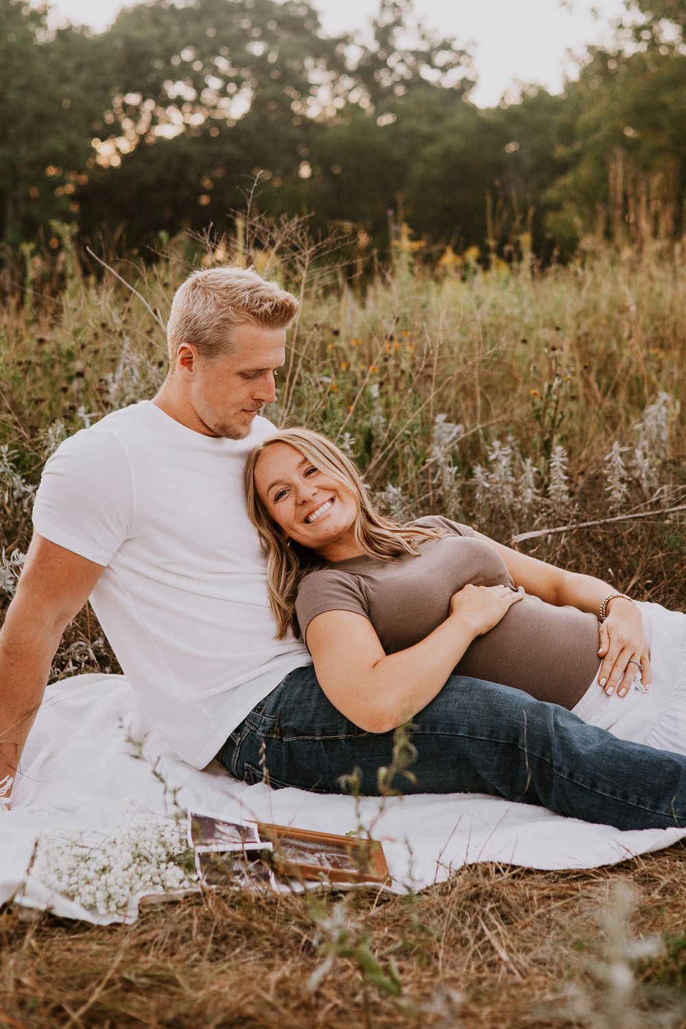 Pregnant couple sitting in a wildflower field, woman resting on man's lap, both smiling warmly during maternity photoshoot.