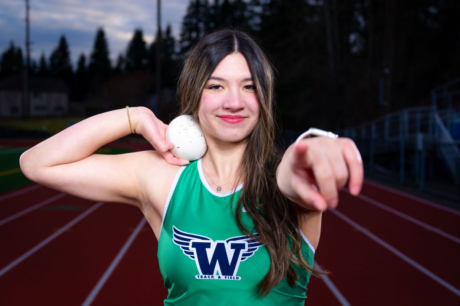 A female track and field athlete from Woodinville High School is posing with a shot put while pointing at the camera.