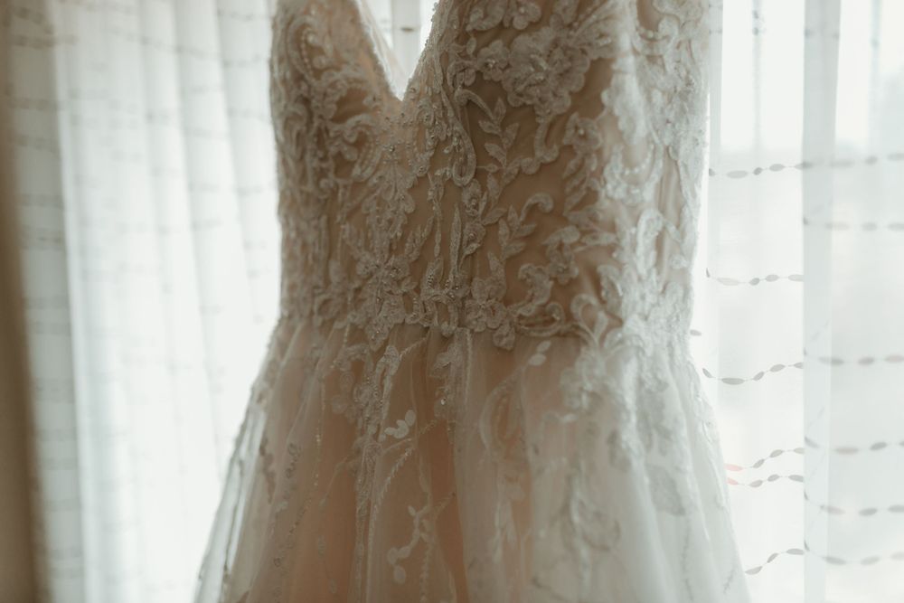 Intricate lace V-neck wedding dress hanging by window with soft natural light.