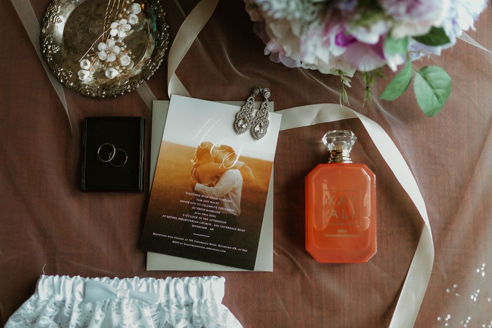 Wedding flat lay with invitation, orange perfume bottle, rings, earrings, and bridal bouquet.