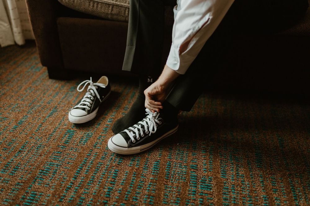Groom putting on black high-top Converse sneakers while sitting on chair before wedding.