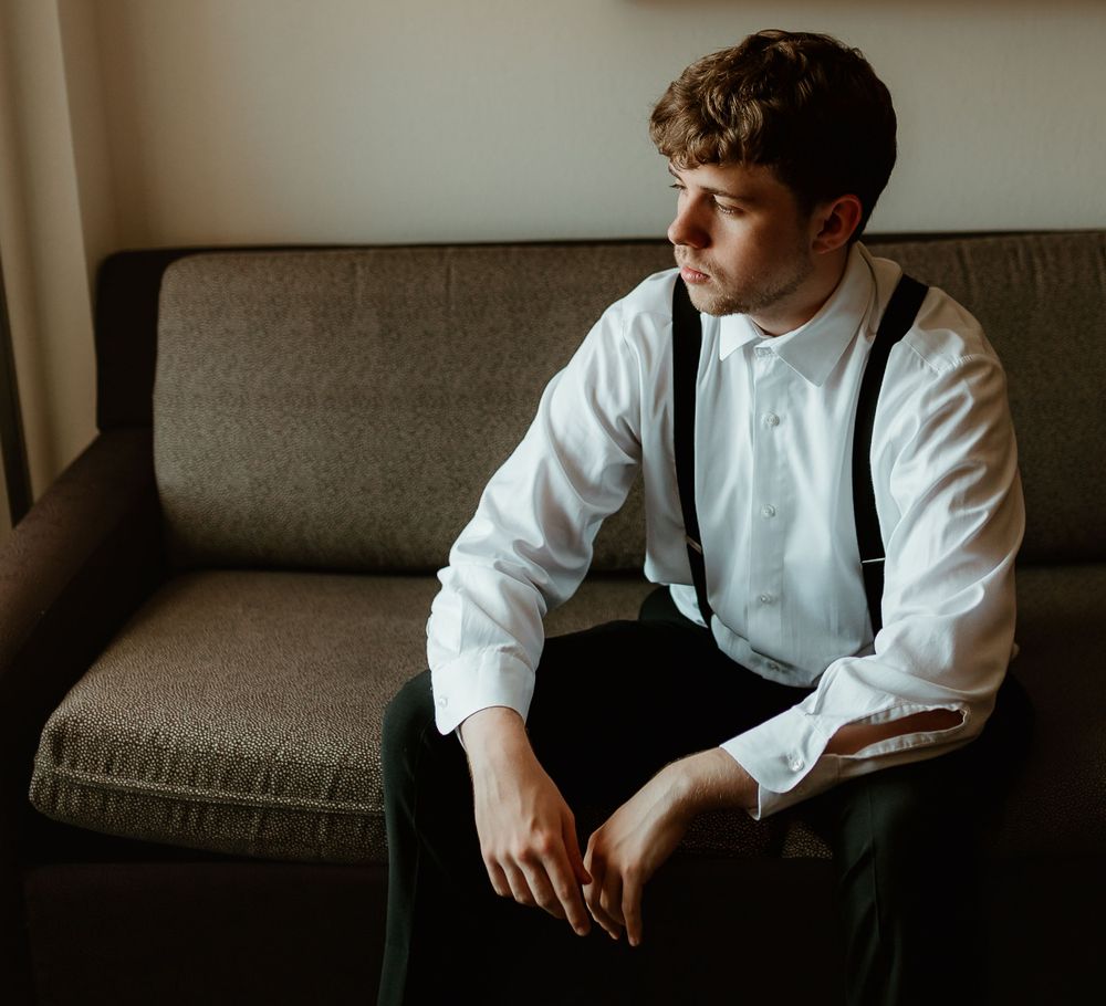 Groom in white shirt and suspenders sitting on couch preparing for wedding ceremony.