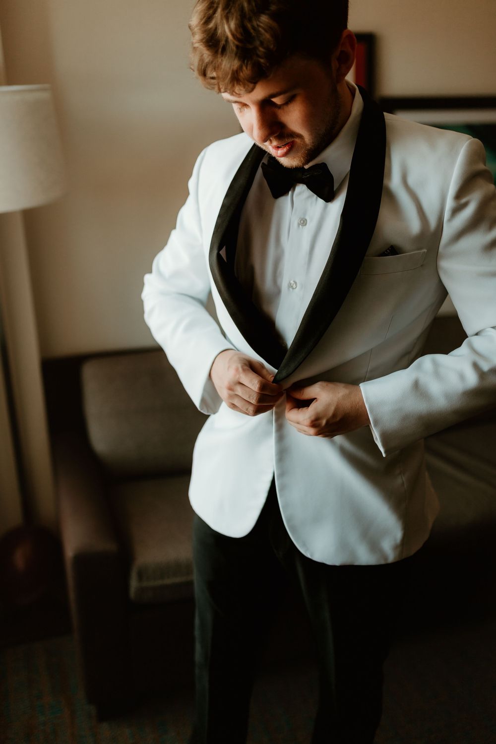 Groom buttoning white tuxedo jacket with black lapels and bow tie before wedding.