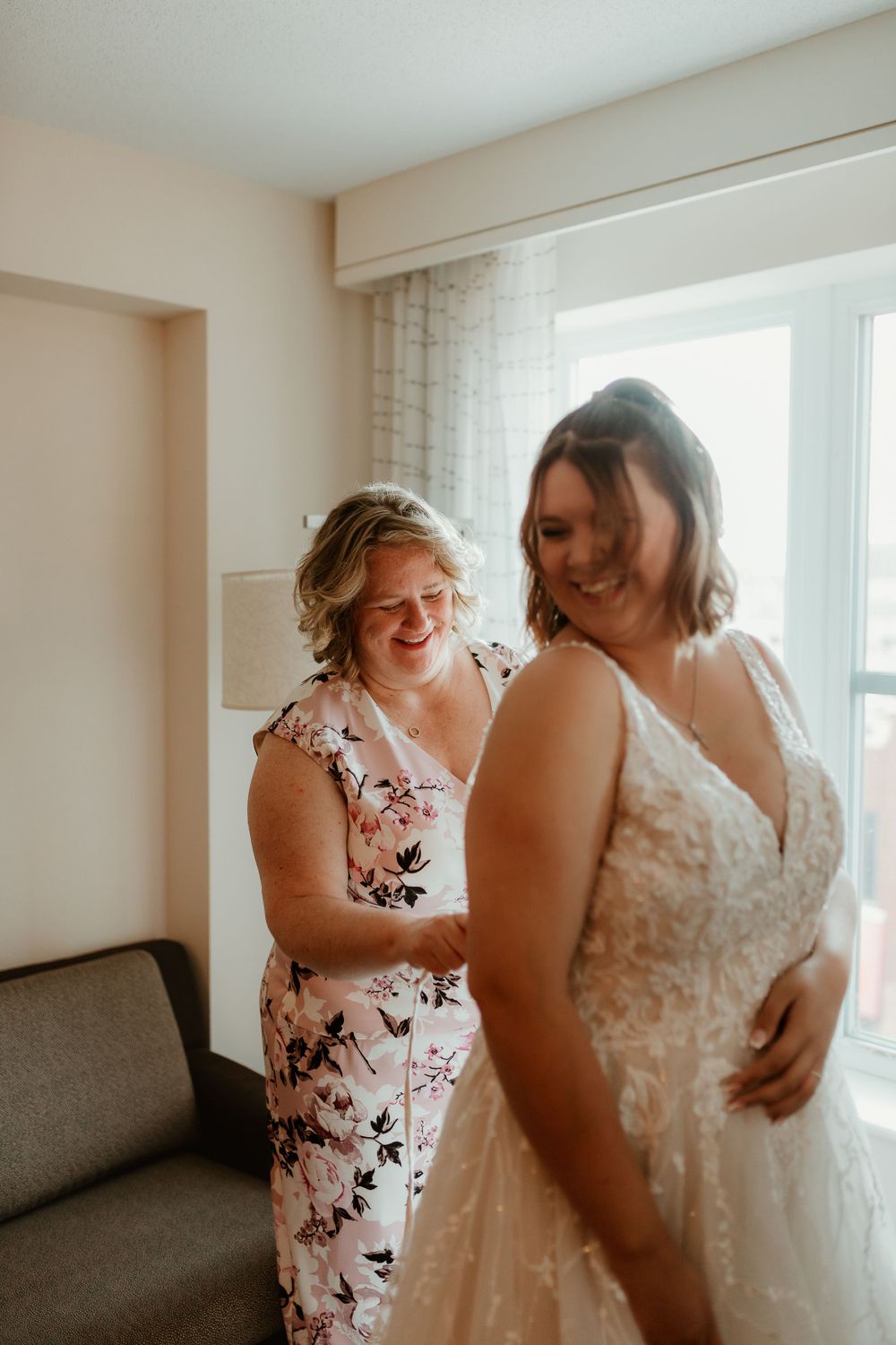A bride in lace wedding gown laughs with a woman in floral dress helping her get ready indoors.
