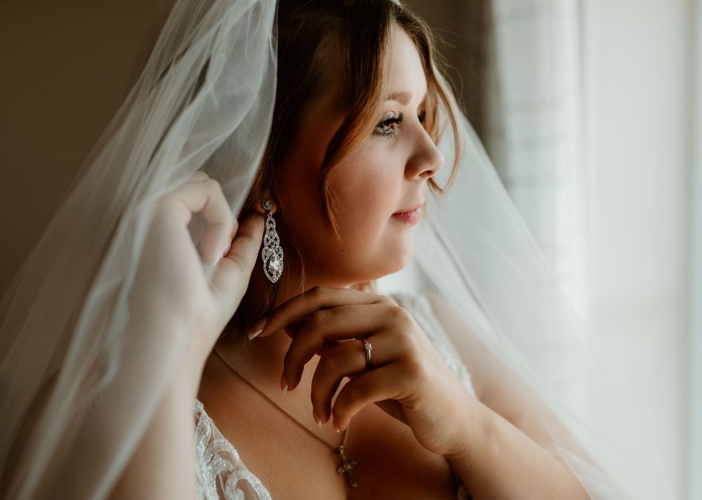 A bride wearing a veil and crystal earrings gazes thoughtfully out a window on her wedding day.
