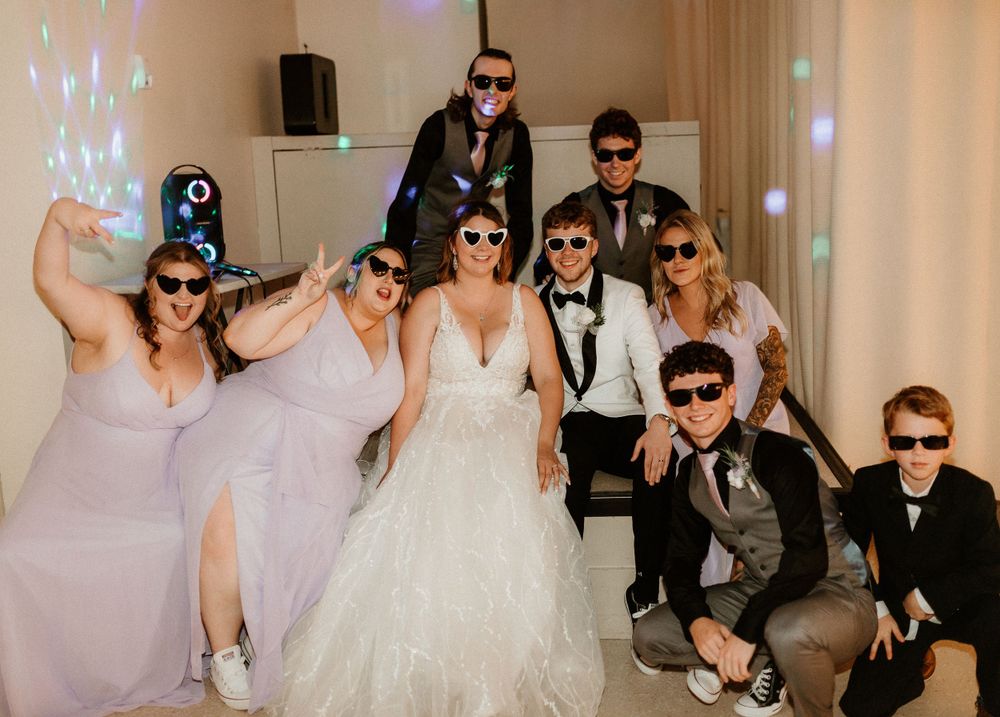 Wedding party posing with sunglasses on dance floor during lively reception celebration.