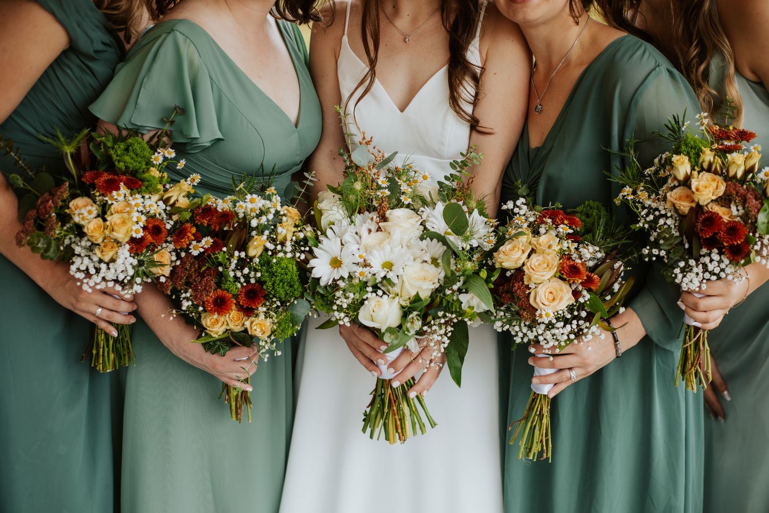 Bride in white gown and bridesmaids in sage green dresses holding colorful wildflower bouquets.