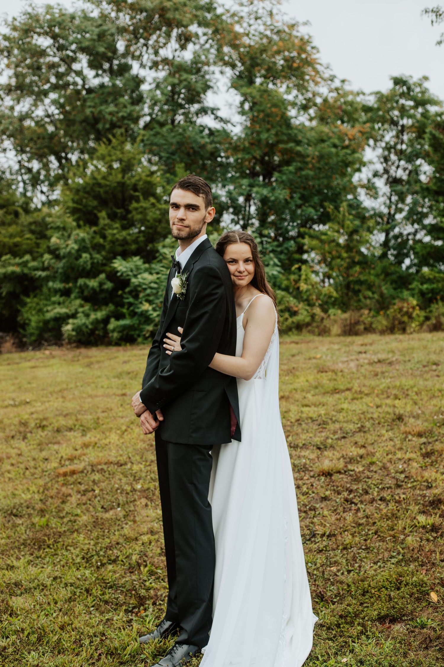 Bride in white gown and groom in black suit pose back-to-back in a green outdoor field on their wedding day.