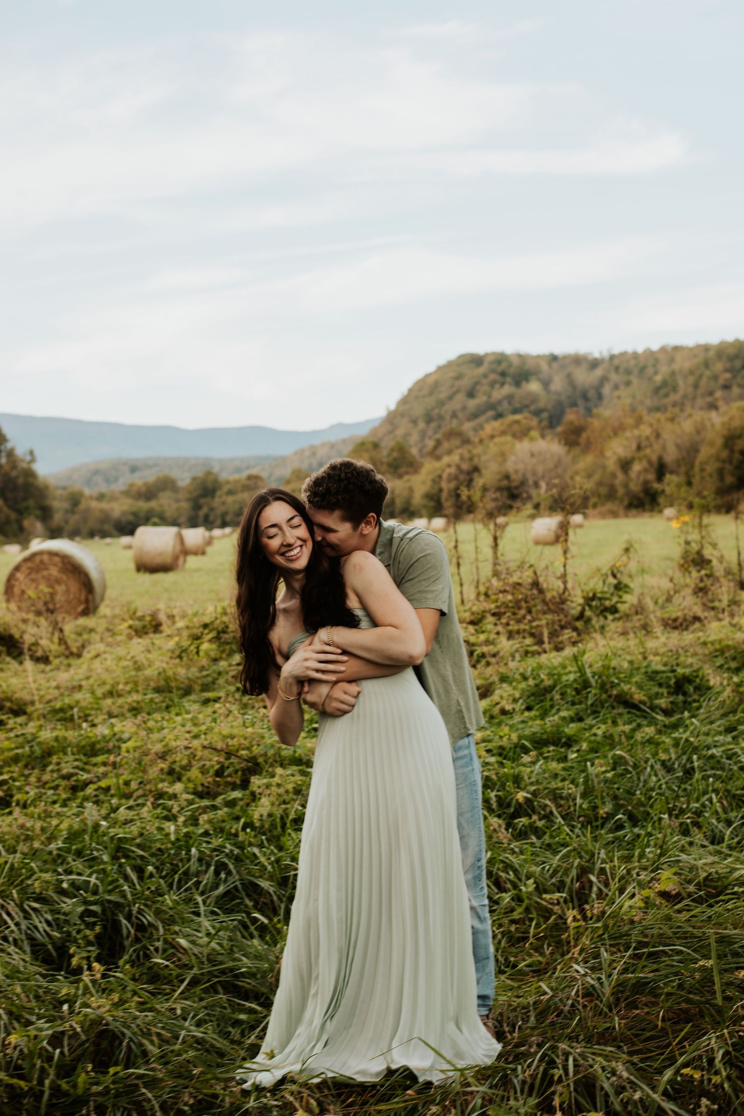 Couple embraces laughing in green field with hay bales and mountain backdrop on cloudy day.