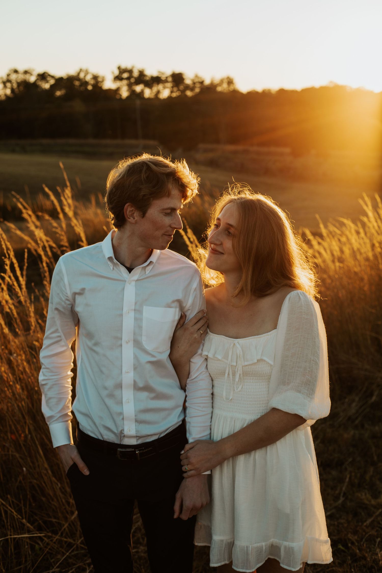 Couple gazes at each other in golden hour sunlight in field, man in white shirt, woman in white dress.