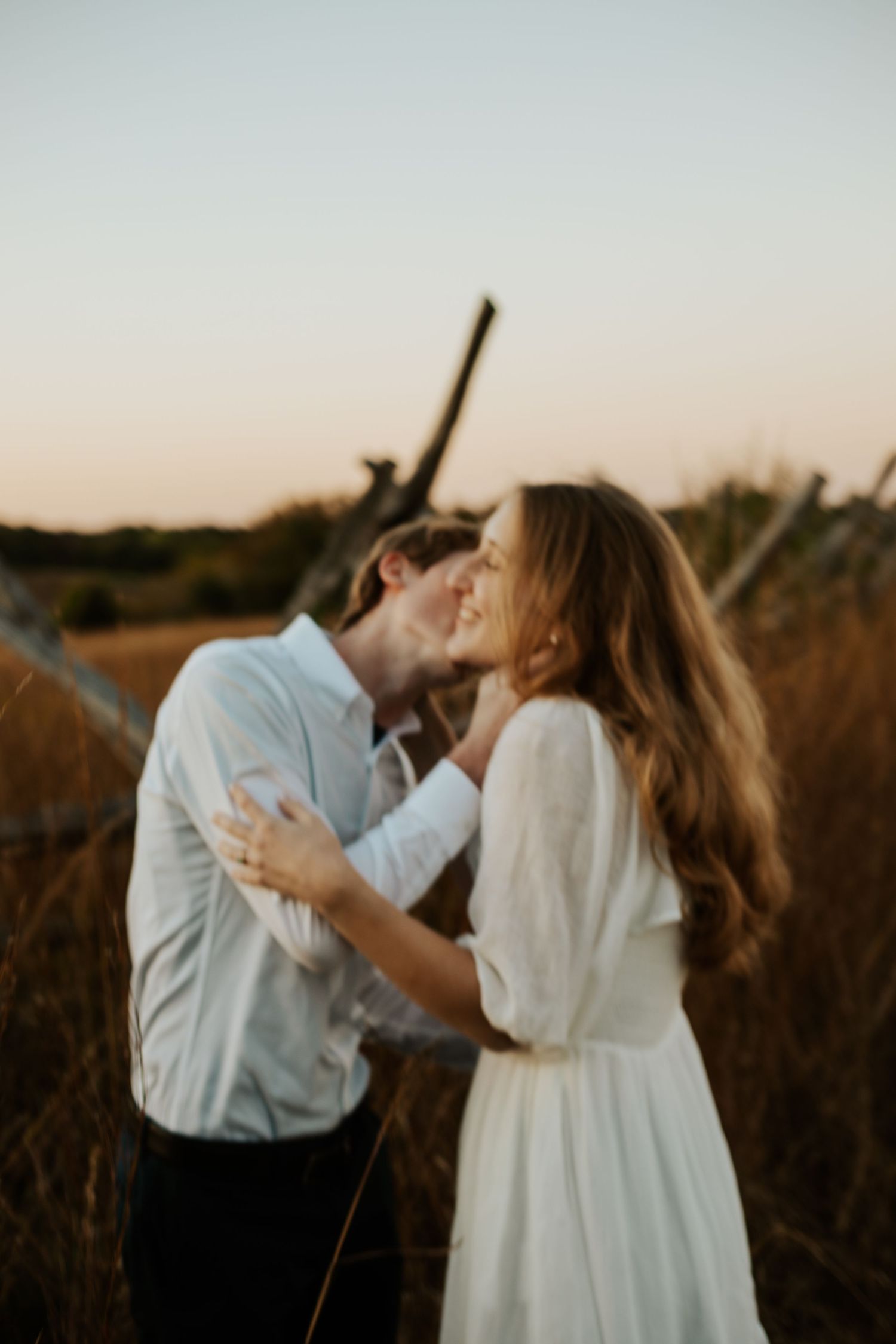 Couple shares tender kiss at golden sunset in tall dry grass field with rustic wood backdrop.