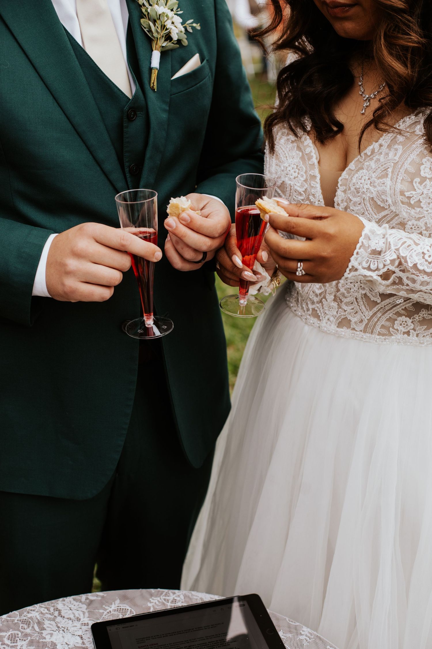 Bride and groom holding red champagne flutes at wedding reception, bride in lace gown, groom in green suit.