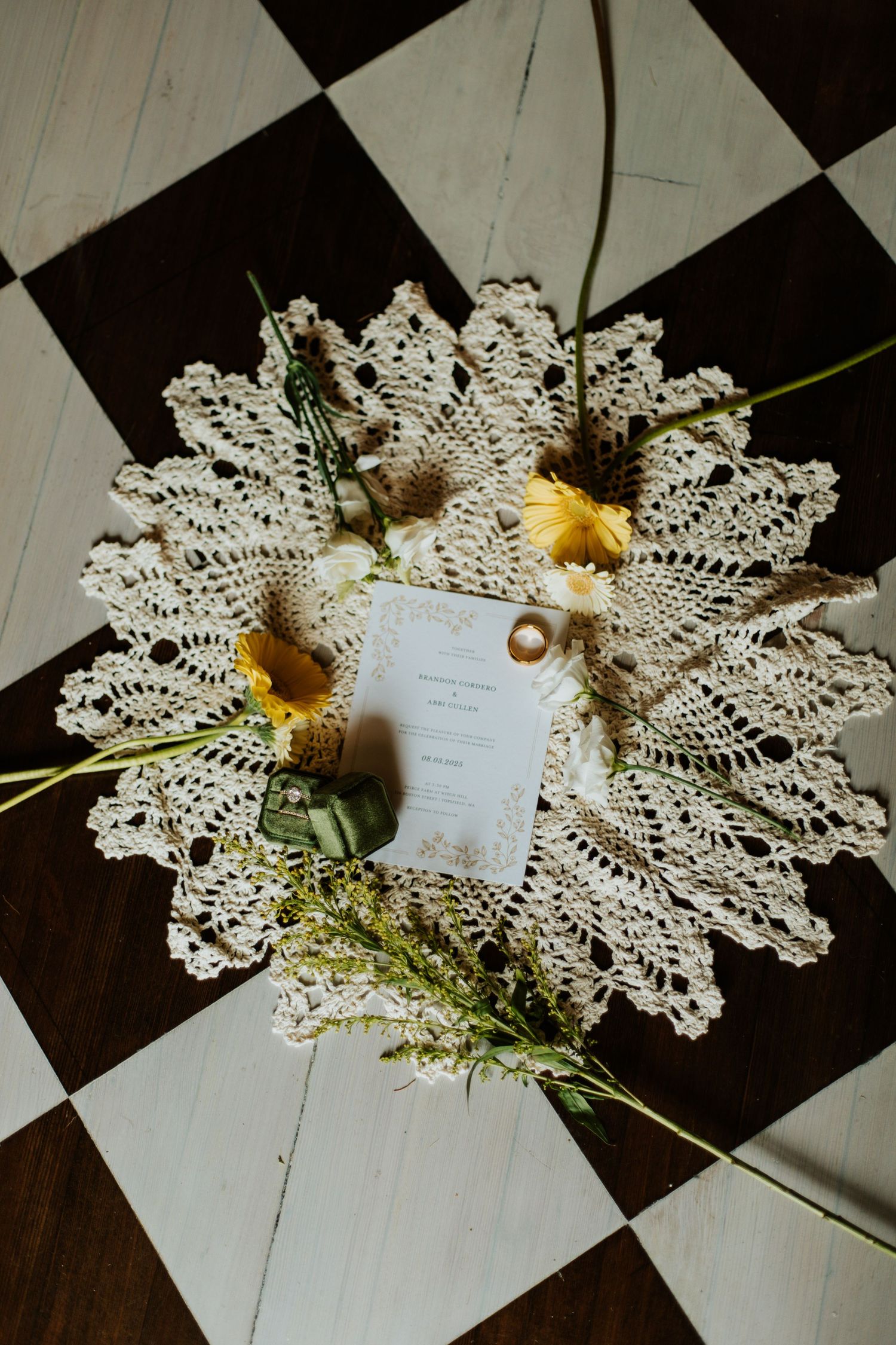 Wedding rings and ceremony card on lace doily with yellow flowers on checkered floor.