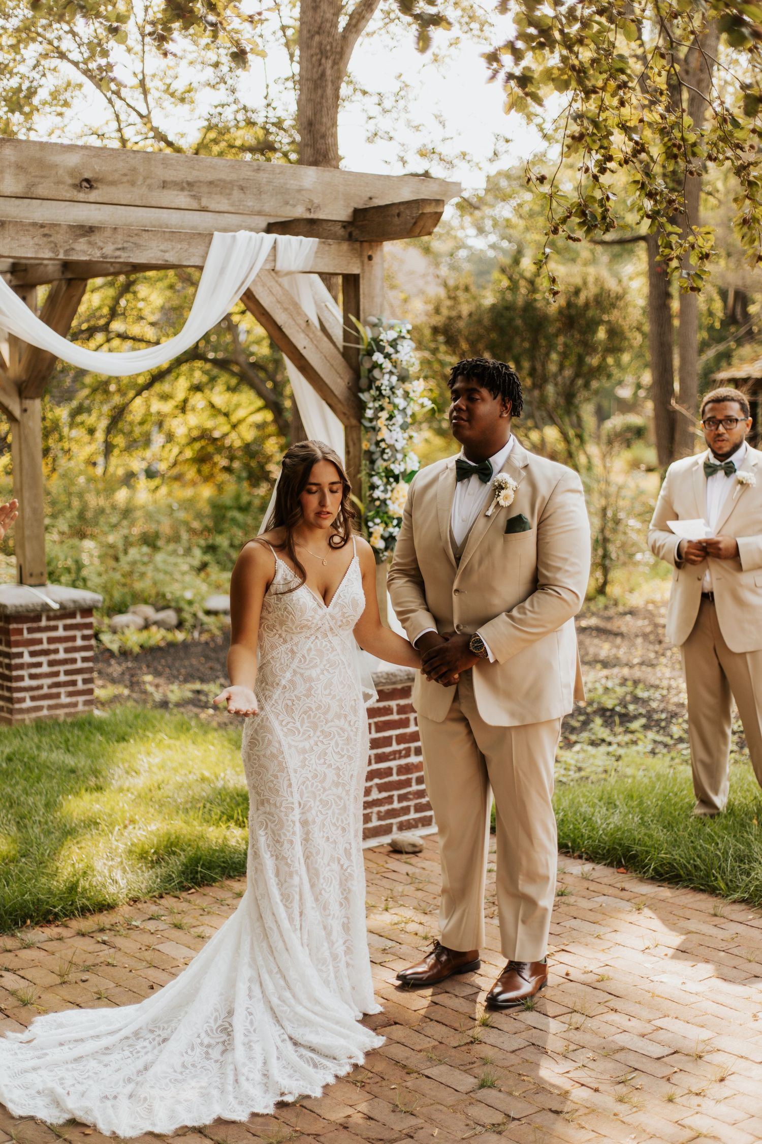 Couple exchanges vows at outdoor wedding ceremony under rustic wooden arch adorned with white draping and floral greenery.