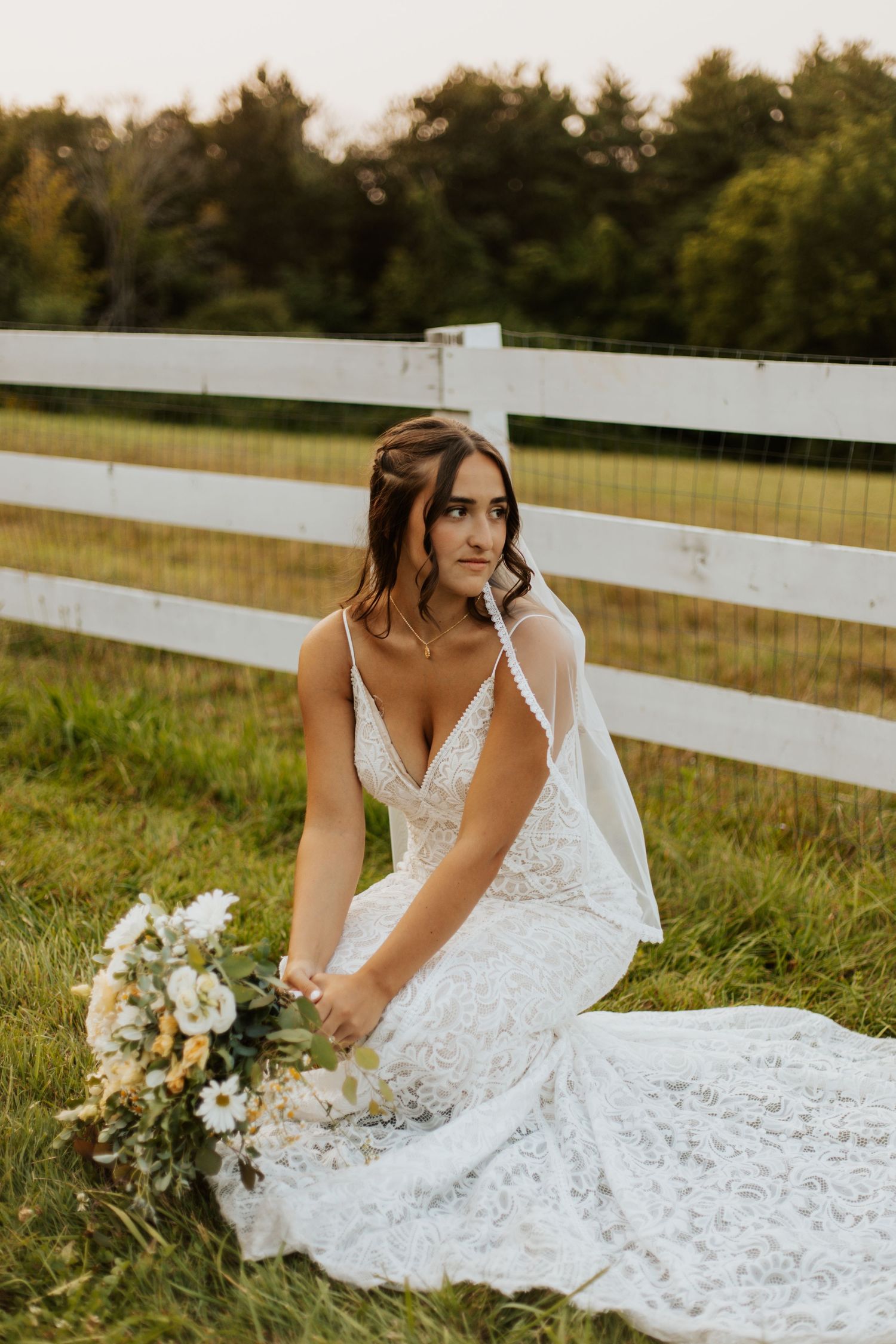 Bride in lace wedding dress sits on grass by white fence holding white and yellow bouquet at sunset.