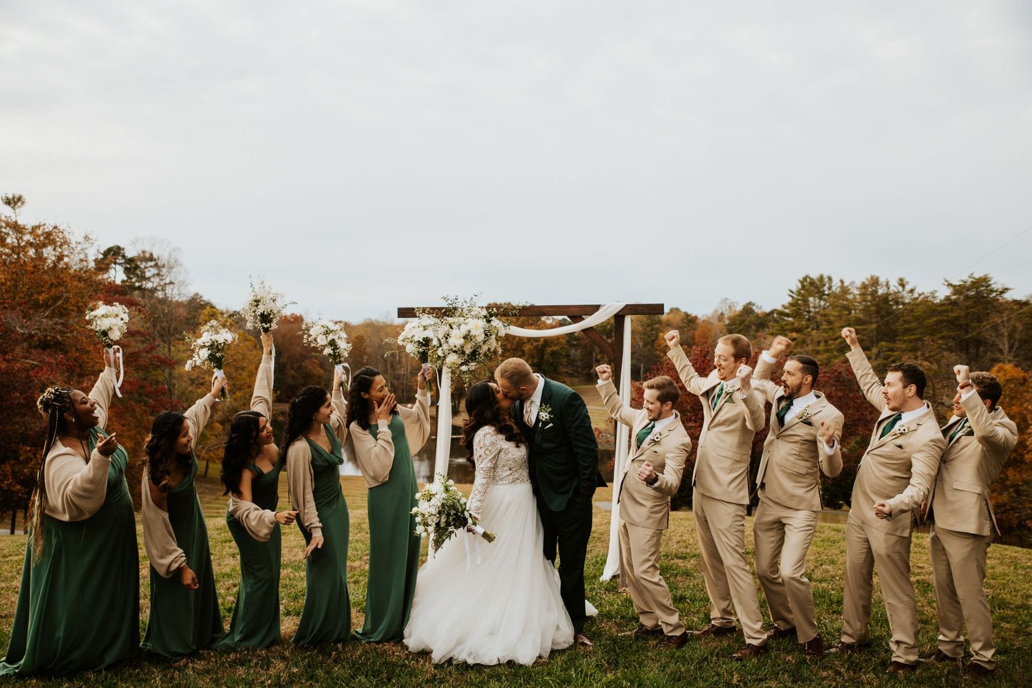 Wedding party cheers as bride and groom kiss under wooden arch at outdoor fall ceremony.