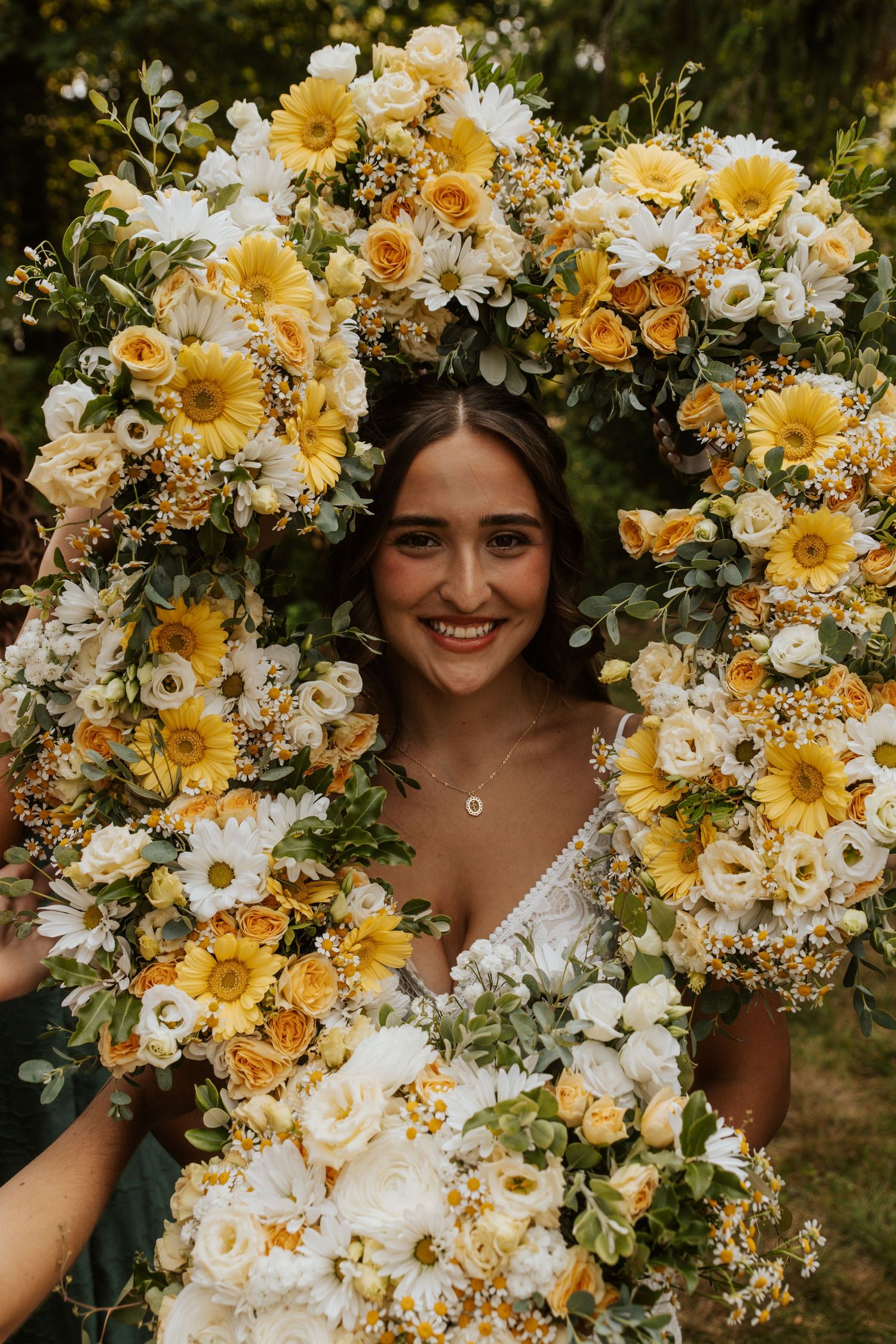 Smiling bride framed by large circular arrangement of yellow sunflowers, white daisies, and roses.