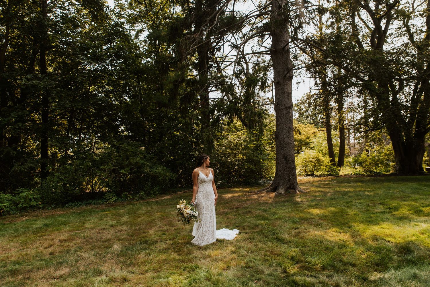Bride in white gown holding bouquet stands on lush green lawn surrounded by tall trees on a sunny day.