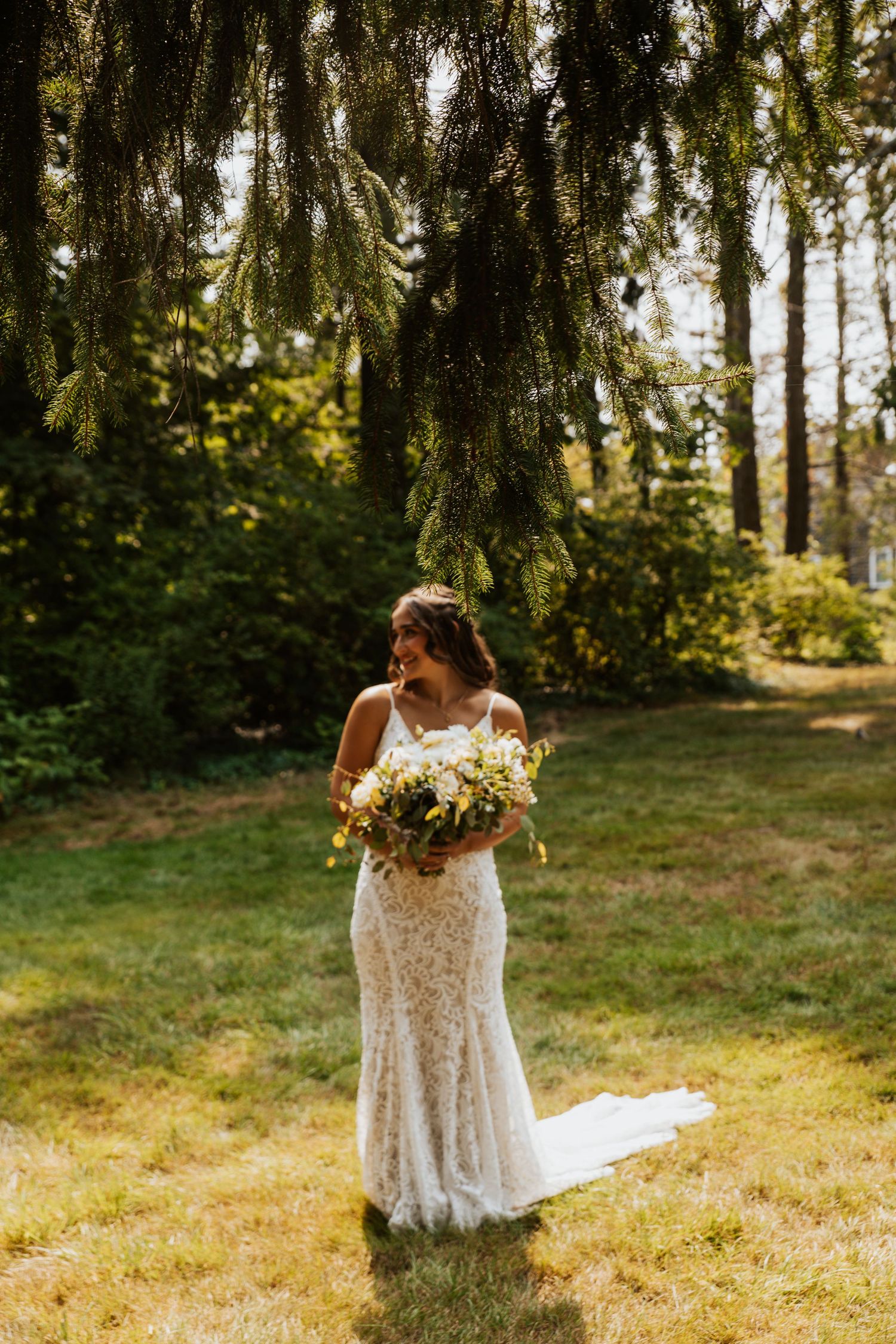 Radiant bride in lace fitted wedding gown holding a wildflower bouquet beneath pine trees outdoors.